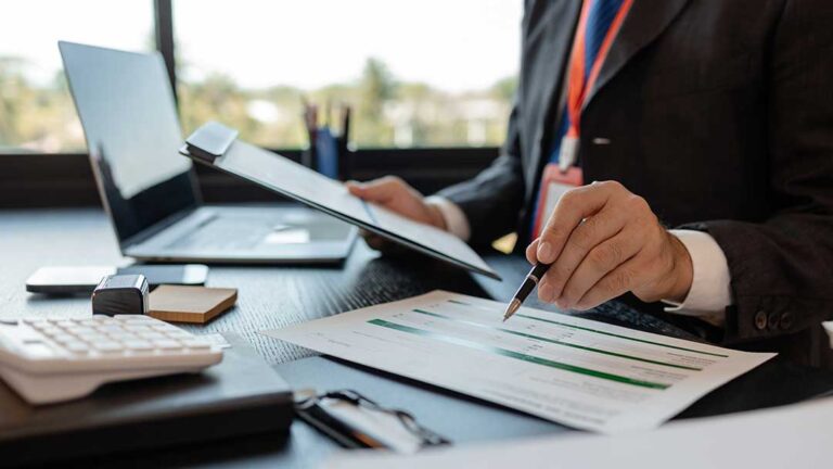 Close-up of a person reviewing a paycheck stub and calculator at a kitchen table with paperwork.